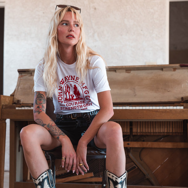 woman sitting and wearing john wayne MFGCO white womans tee with desert scenery and cowboy 