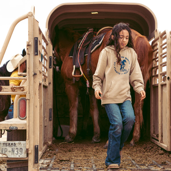 girl walking off horse trailer with hoodie on that has john wayne on a bucking horse and "john wayne stock and supply co." bordering it design