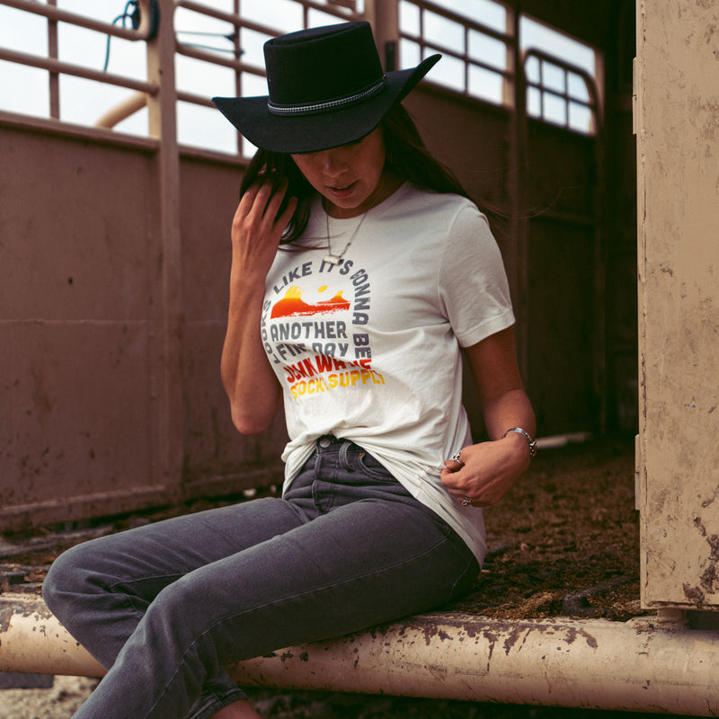 woman sitting wearing t-shirt with "looks like it's gonna be another fine day john wayne stock & supply" and desert scene on it