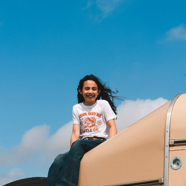 girl sitting wearing white t-shirt with "john wayne cattle co." and cow design