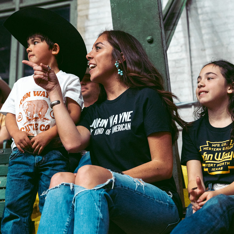 woman sitting with children and wearing black womans tee with "john wayne my kind of american" in white 