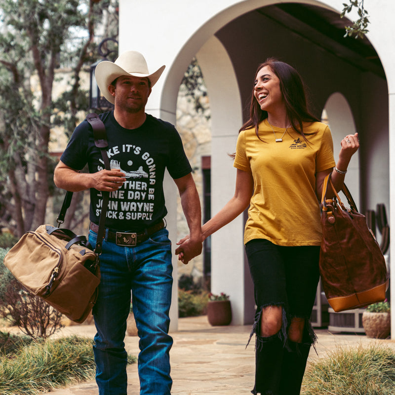 man and woman holding hands with man wearing front of black t-shirt with "looks like it's gonna be another fine day john wayne stock & supply" and desert scene on it