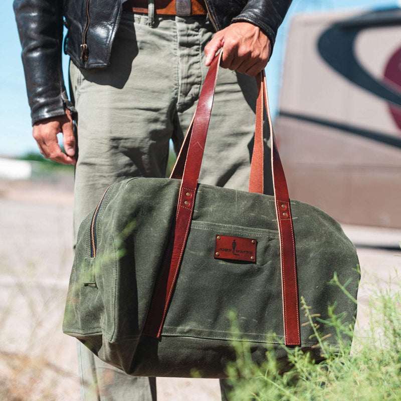 man holding green waxed canvas duffle bag with leather handles and john wayne silhouette patch