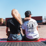 back of man sitting next to woman and wearing back of man wearing john wayne MFGCO pigment white tee with desert scenery and cowboy 